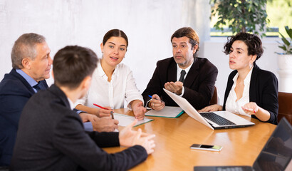 Focused young and experienced business partners sitting at table in office meeting room, discussing projects and ideas with interest