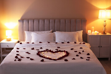 Heart made with beautiful rose petals and burning candles on bed indoors