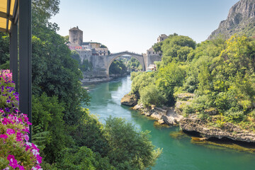 Fototapeta premium The Stari Most bridge, old bridge on Neretva river in Mostar town, Bosnia and Herzegovina, Europe. Old bridge arches across a river, flanked by historic buildings and lush greenery on a sunny day.