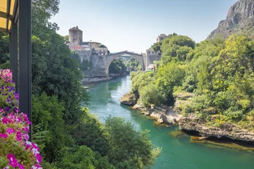 The Stari Most bridge, old bridge on Neretva river in Mostar town, Bosnia and Herzegovina, Europe. Old bridge arches across a river, flanked by historic buildings and lush greenery on a sunny day.