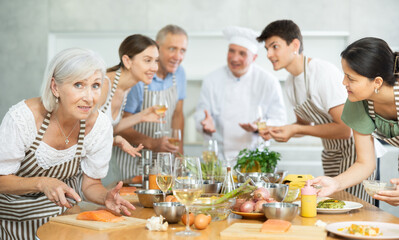 Smiling enthusiastic elderly woman participating group culinary class, engrossed in cooking process mirroring chef instructor steps..