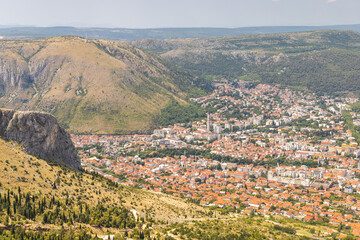 Fototapeta premium View of Mostar from the Fortica hill, Bosnia and Herzegovina, Europe. Scenic overlook of a city nestled in a valley, showing the urban architecture surrounded by hills and mountains.