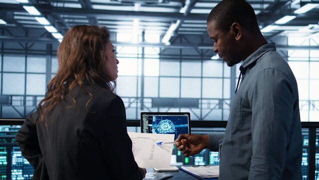 Data center team reviewing paperwork in server room, analyzing system reports using AI on tablet. Artificial intelligence server room admins looking over financial documents, camera B