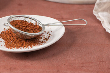 Fine cocoa powder in a metal sieve on a white plate, with some powder spilled around, set against a brown background