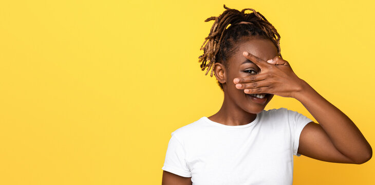 Happy young black woman closing her face with one palm, looking with one eye through her fingers at camera, yellow studio background, empty space. Cheerful african american lady playing hide and seek