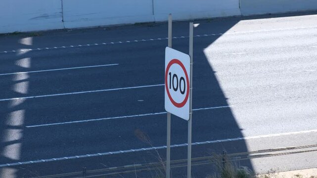 A speed limit sign displaying "100 km/h" stands in the middle of an Australian highway or motorway while passing vehicles or high-speed traffic flow by. Melbourne, Victoria, Australia.