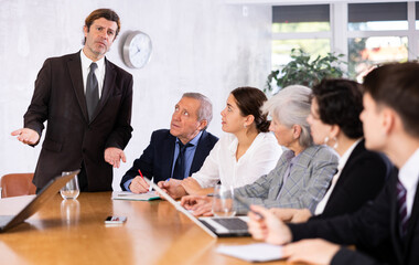 Displeased male manager scolding at young, adult, mature office workers sitting with reports and laptops at table in meeting room