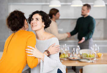 Fototapeta premium Women hug each other and kiss on the cheek while greeting each other before a festive dinner. Men shaking hands in the background