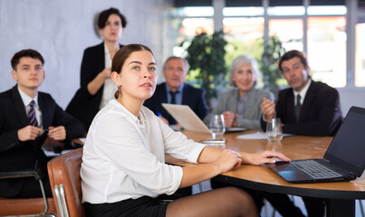 Portrait of positive young female office worker sitting at table with colleagues, listening with interest and watching presentation during business meeting
