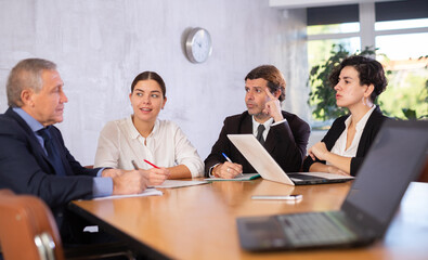Focused young and experienced business partners sitting at table in office meeting room, discussing projects and ideas with interest