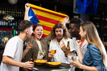 Group of Spain football team fans spending time in bar, drinking bear and having fun. People with Estelada flag in pub.