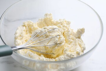 Bowl with whisk and whipped cream on white marble table, closeup