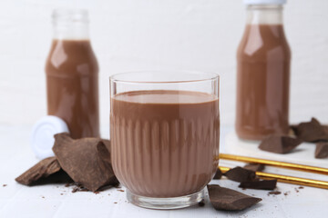 Tasty chocolate milk in glass and pieces of chocolate on white tiled table, closeup