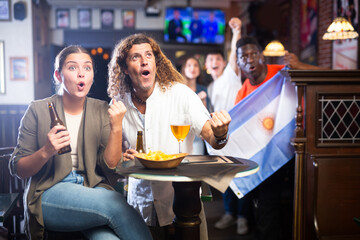 Expressive young adult couple sitting at table in sports bar, drinking beer with snacks and emotionally rooting for favorite Argentinean team while watching football match