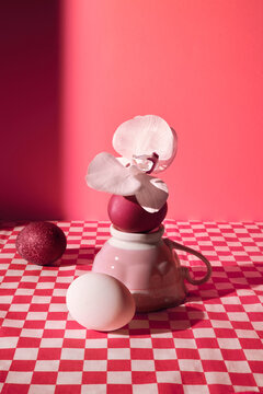 Abstract still life with pink cup, eggs and flower on checkered table.