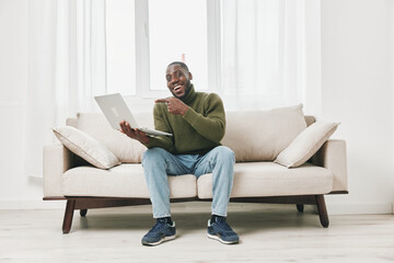 Happy young man working on a laptop at home, wearing a green sweater and jeans, sitting on a light...