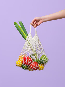 Hand Holding Mesh Bag with Vegetables on Light Purple Background