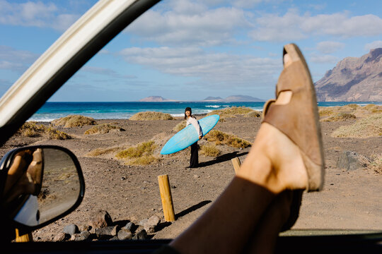 Asian woman stands on the beach seen through the open window of a car