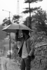 young man standing roadside holding an umbrella on a rainy day, wearing a jacket over shirt and cargo pants.