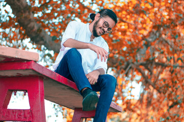 A young man sits on a red bench beneath an autumn tree with vibrant orange leaves laughing, holding...