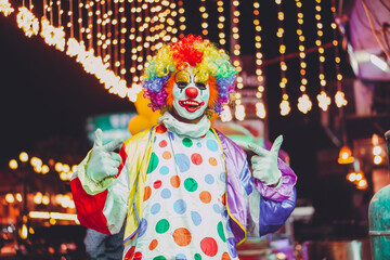 a colorful clown with a wig and polka-dot outfit, doing a happy hand gesture against a festive backdrop of decorative lights.