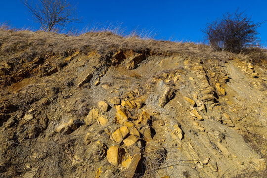 The photograph depicts a hillside with exposed earth and rocks. The earth is brown-yellow in color, and the rocks have a yellow tint, creating a contrast. On the top of the hill, dry grass and bushes 