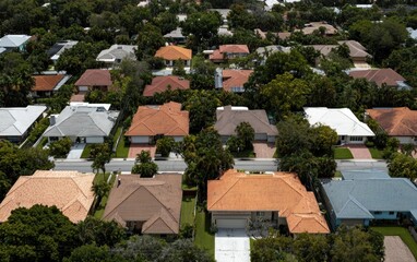 Fototapeta premium Aerial view of a suburban neighborhood with houses featuring a variety of roof colors and styles. Lush green trees are interspersed between the homes. The image offers a bird's-eye perspective of a