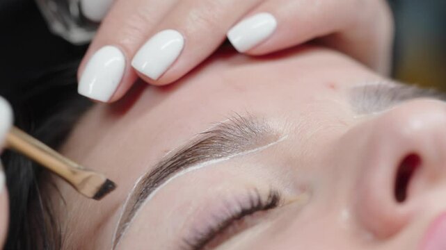 Beautician applies eyebrow tint with a precision brush while the client lies relaxed with closed eyes during a brow shaping and coloring treatment.
