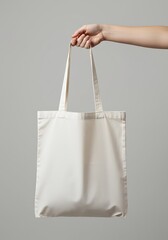 A hand gently holds a simple, elegant cream-colored tote bag against a muted gray backdrop, showcasing its versatility and understated charm.