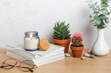 Burning candle, books, eyeglasses, plants and vase creating cozy atmosphere on wooden desk