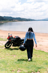 Obraz premium Young woman biker is walking away from her motorcycle parked near a lake, carrying her helmet