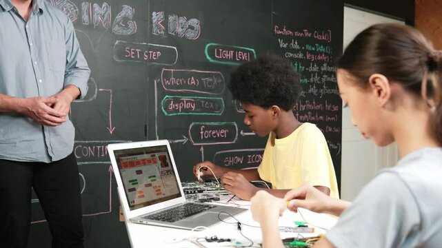 Teacher teaching about coding system while diverse academic student fixing main board at table with laptop display programming prompt or engineering code at STEM technology classroom. Edification