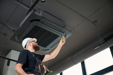 Bearded man staying on ladder installing air conditioner at the office. Ceiling mounted cassette...