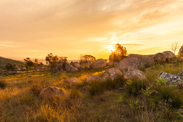 A backlit landscape of the sun setting in a cloudy sky over granite boulders and rocks strewn over grasslands and amongst trees in granite country near Tenterfield in New South Wales, Australia.