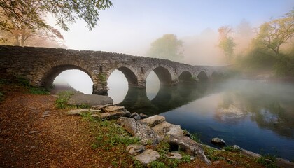 Fototapeta premium Atmospheric ancient stone bridge emerging from the fog over a serene river reflecting mystery