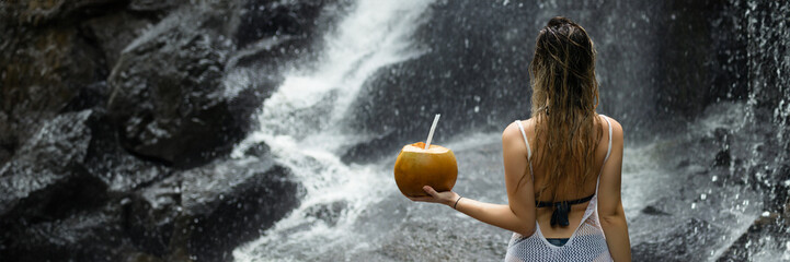 Beautiful sexy woman stands facing a waterfall in Bali and holds a coconut in her hand