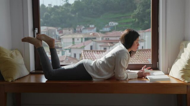 Woman lying by her window reading a book.