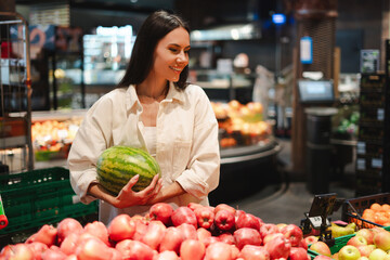 Smiling, attractive Latin woman choosing watermelon and apples in supermarket