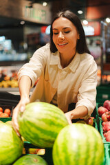 Fototapeta premium Smiling woman choosing watermelon in supermarket fruit section