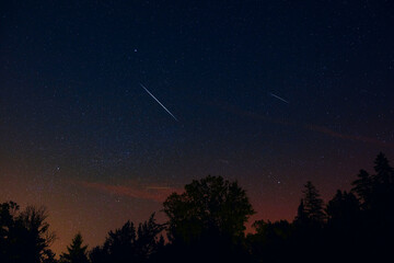 Milky Way stars with meteor shower trails and countryside tree silhouettes.