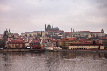 Panoramic view of Prague with historic buildings, red rooftops, and Prague Castle in the background on a cloudy day.