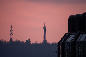 Obraz premium Silhouettes of Petřín Lookout Tower, St. Lawrence Church and a telecom mast in Prague at sunset, set against a soft pink sky with modern architecture in the foreground.