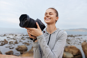 Young woman photographer enjoying nature with camera near the sea, smiling and feeling joyful, capturing beautiful moments outdoors