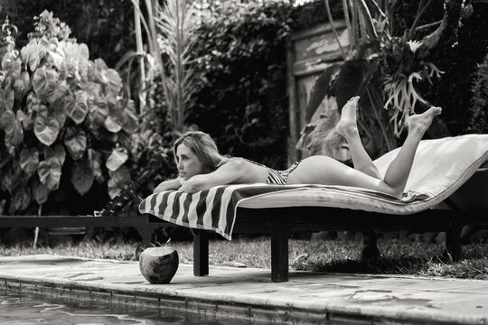 Woman relaxing on sunlounger near pool, drinking young coconut