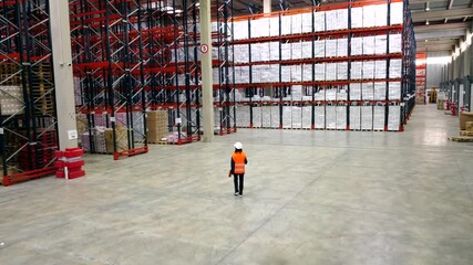 Professional logistics supervisor wearing high visibility safety vest and hardhat walking confidently through spacious warehouse, checking inventory and monitoring distribution center operations
