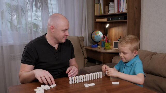 Cheerful father and son play with domino bones sitting at a wooden table, they put dominoes in a row. 