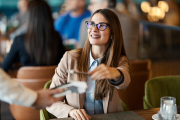 A young happy woman is smiling at waiter and paying her bill with e-banking mobile app.