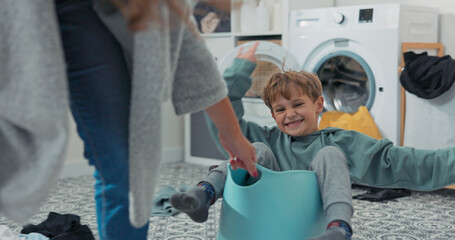 A cheerful son plays in the laundry room, sitting inside a bowl of clothes. His mother gently pulls...
