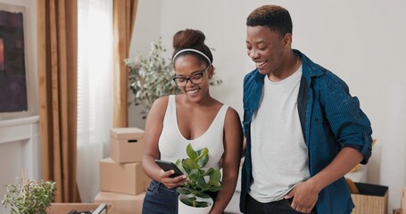 In their fresh apartment, a couple poses for a selfie. The woman holds a potted plant while smiling with her partner, capturing and sharing the excitement of their move online.