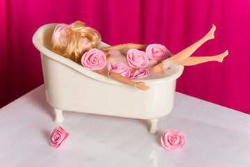 Top view doll in bath with soap foam and pink flowers on a pink background.